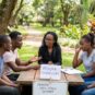 A female counselor with braided hair and glasses leading a grief support group for four young friends at a wooden outdoor table in a lush park setting