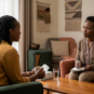 A compassionate female counselling psychologist with short natural hair listens attentively to a young woman client during a confidential therapy session in a warm, inviting modern office.