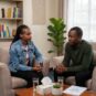 A recognizable female African counselling psychologist in her denim jacket and red lipstick listens with profound empathy to a distressed yet open young male client during a confidential therapy session in a warm, professional Nairobi office featuring natural lighting, bookshelves with varied literature, subtle African art, comfortable armchairs, a potted plant, and essential details on a low table.