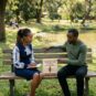 An expert female African counselling psychologist in a blue and patterned dress sitting on an outdoor park bench, calmly taking notes while guiding a distressed male client. Between them on the bench is a wooden sign that reads "Reclaiming Your Space: Setting Healthy Boundaries" with a small picket fence icon, set in a serene, natural Nairobi park environment with trees and a pond in the background.